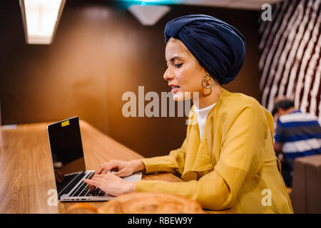 Un giovane malese donna asiatica è la digitazione e lavorare o studiare sul suo computer portatile in un caffè durante la giornata. Indossa un turbante hijab. Foto Stock