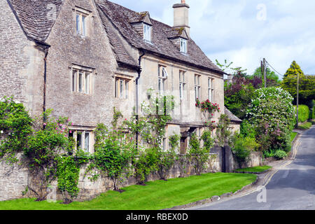 Villaggio strada lungo il vecchio cottage inglese nelle zone rurali del Cotswold campagna, circondato da fiori, arbusti, alberi del giardino, su una soleggiata giornata estiva . Foto Stock