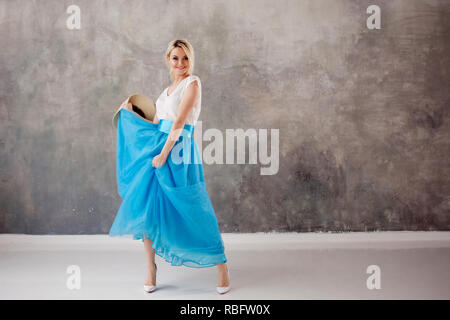 Bella giovane donna in un lussureggiante mantello blu, vorticoso e balli. Estate stile, cappello di paglia con nastro nero. Grigio sfondo testurizzata Foto Stock