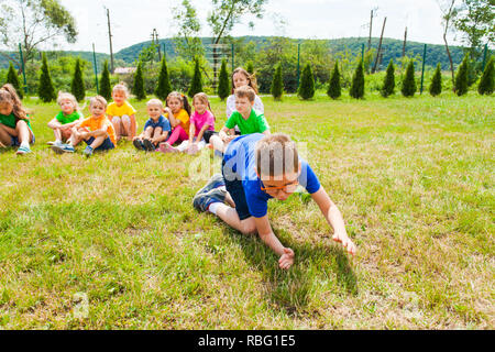 Scolaro in bicchieri crowling inginocchiato sul prato davanti i suoi compagni, seduto su un terreno all'aperto in estate park. Lezioni sulla recitazione skil Foto Stock