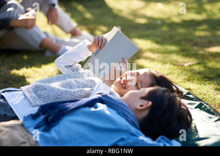 Due piccoli bambini asiatici un ragazzo e una ragazza che giace sul retro su erba nel parco la lettura di un libro insieme con i genitori seduti in background. Foto Stock