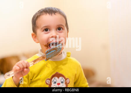 Bambino felice lambisce un cucchiaio con il cioccolato. Felice ragazzo di mangiare una torta al cioccolato. Funny baby mangiare il cioccolato con un cucchiaio. Foto Stock