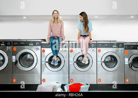 Due amiche allegro in attesa per il lavaggio seduti sulle macchine di lavaggio nel servizio di lavanderia self-service Foto Stock