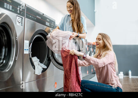 Due amiche allegro divertimento caricando i vestiti nella macchina di lavaggio nel servizio di lavanderia self-service Foto Stock