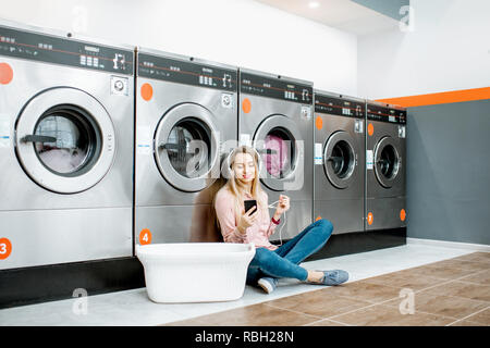 Giovane donna in attesa per i panni da lavare seduto sul pavimento e ascolto di musica presso il servizio di lavanderia self-service Foto Stock