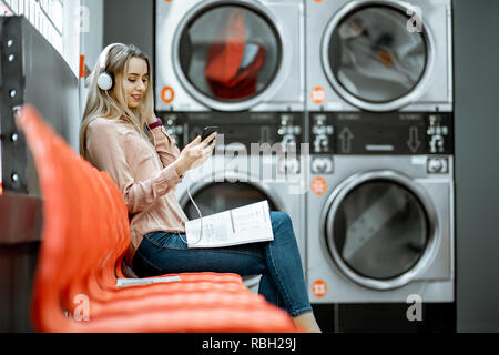 Giovane donna godendo la musica di attesa per i panni da lavare seduto sulla sedia al servizio di lavanderia self-service Foto Stock