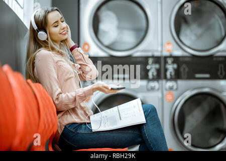 Giovane donna godendo la musica di attesa per i panni da lavare seduto sulla sedia al servizio di lavanderia self-service Foto Stock