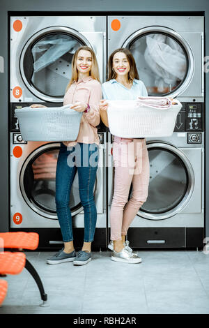 Due giovani donne in piedi insieme con ceste piene di capi in lavanderia con asciugatrice macchine sullo sfondo Foto Stock