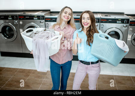 Ritratto di un allegro due amiche in piedi insieme con ceste piene di panni da lavare in lavanderia self-service Foto Stock