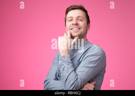 Fiducioso giovane uomo bello in maglietta blu tenere le braccia incrociate e sorridere mentre in piedi contro sfondo rosa Foto Stock