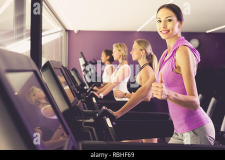 Sorridente montare le ragazze in esecuzione sul tapis roulant nel centro fitness dando pollice in alto Foto Stock