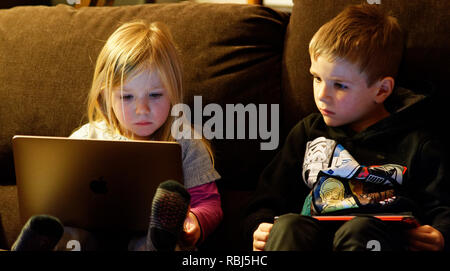 Una bambina di 4 anni seduto su un divano con un computer portatile mentre suo fratello (6 anni) orologi. Foto Stock