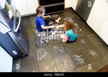 Due bambini che utilizzano i gessi per disegnare su un pavimento di ardesia nella loro cucina Foto Stock