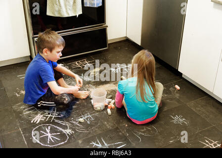 Due bambini che utilizzano i gessi per disegnare su un pavimento di ardesia nella loro cucina Foto Stock