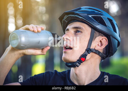 Un ciclista in un casco di bere acqua da una bottiglia mentre passeggiate a cavallo nel parco tra gli alberi. Concetto di attivo e uno stile di vita sano Foto Stock