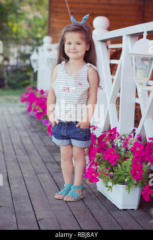 Bellissima ed elegante simpatico baby girl Brunette con capelli in posa su legno giardino pieno di fiori che indossa jeans minuscole magliette e ariosa skivy underwaist e b Foto Stock