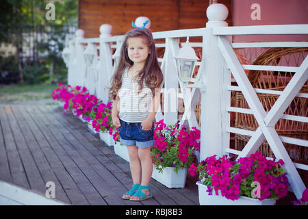 Bellissima ed elegante simpatico baby girl Brunette con capelli in posa su legno giardino pieno di fiori che indossa jeans minuscole magliette e ariosa skivy underwaist e b Foto Stock