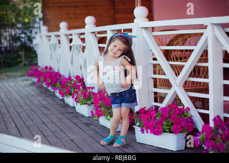 Bellissima ed elegante simpatico baby girl Brunette con capelli in posa su legno giardino pieno di fiori che indossa jeans minuscole magliette e ariosa skivy underwaist e b Foto Stock