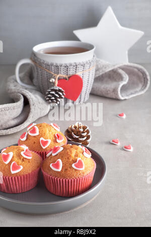 Muffin decorati con cuori di zucchero e una tazza con cuore rosso su sfondo grigio chiaro. Il giorno di San Valentino o in inverno il concetto di compleanno. Foto Stock