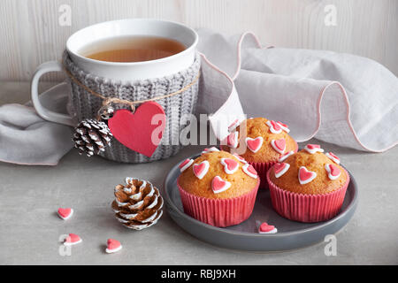 Muffin decorati con cuori di zucchero e una tazza con cuore rosso su sfondo grigio chiaro. Il giorno di San Valentino o in inverno il concetto di compleanno. Foto Stock