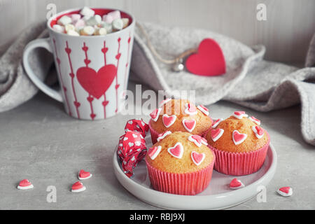 Muffin decorati con cuori di zucchero e una tazza con cuore rosso su sfondo grigio chiaro. Il giorno di San Valentino o in inverno il concetto di compleanno. Foto Stock