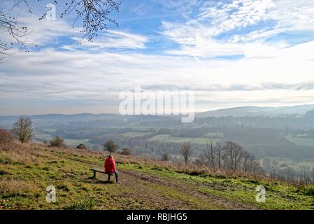 Il camminatore femmina in rosso cappotto seduta sul banco di lavoro guardando a vista dal Comune Ranmore Surrey Hills Dorking su un soleggiato inverni giorno,Surrey in Inghilterra REGNO UNITO Foto Stock