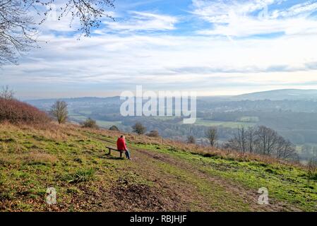 Il camminatore femmina in rosso cappotto seduta sul banco di lavoro guardando a vista dal Comune Ranmore Surrey Hills Dorking su un soleggiato inverni giorno,Surrey in Inghilterra REGNO UNITO Foto Stock