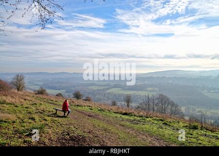 Il camminatore femmina in rosso cappotto seduta sul banco di lavoro guardando a vista dal Comune Ranmore Surrey Hills Dorking su un soleggiato inverni giorno,Surrey in Inghilterra REGNO UNITO Foto Stock