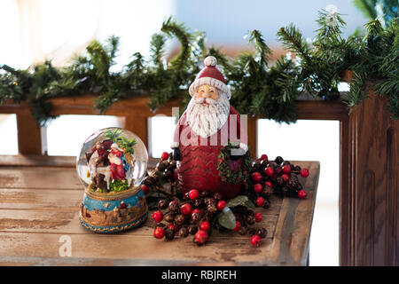 Decorazioni di Natale, una palla di neve con la scena della Natività e Babbo Natale su un tavolo di legno con una ghirlanda su una ringhiera. Foto Stock