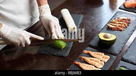 Vista ravvicinata del processo di preparazione di delizioso sushi di laminazione nel ristorante. Mani femminili in guanti monouso per affettare avocado su tavola di legno con th Foto Stock