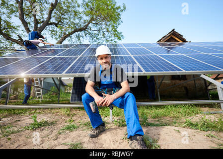 Ritratto maschile di ingegnere tecnico con cacciavite elettrico cercando nella fotocamera seduto di fronte a quasi finito esterna elevata pannello solare foto Foto Stock