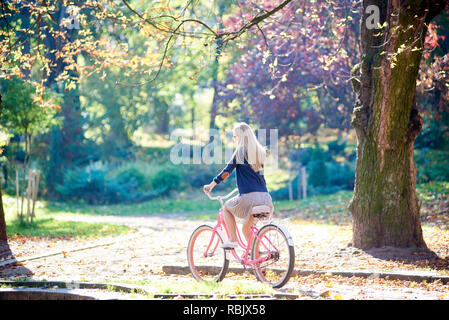 Vista laterale di giovani attiva bionda con i capelli lunghi donna in gonna e camicetta ciclismo pink lady in bicicletta lungo il parco vicolo pavimentato con caduto foglie d'oro su bl Foto Stock