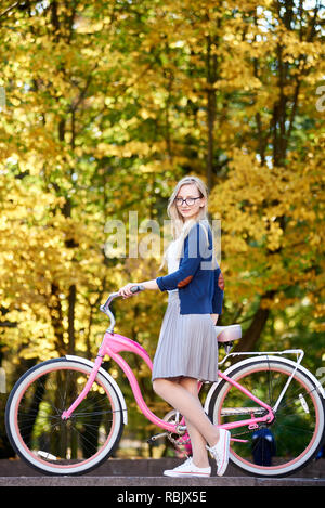 Active bionda dai lunghi capelli ragazza attraente in bicchieri, gonna e camicetta holding gestisce delle moderne pink lady bicicletta su illuminata dal sole autunnale luminose colorate Foto Stock