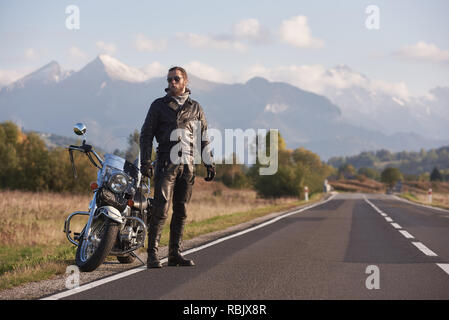 Barbuto tall athletic biker in occhiali da sole scuri, nero abbigliamento in pelle permanente al moderni potenti moto sullo sfondo del verde paesaggio rurale, s Foto Stock