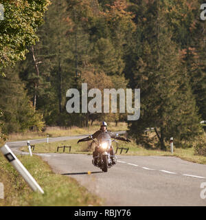 Barbuto motociclista nel casco, occhiali da sole e pelle nera di equitazione abbigliamento moto con stese di bracci lungo vuoto strada asfaltata avvolgimento tra alti alberi verdi sul luminoso giorno d'estate. Foto Stock