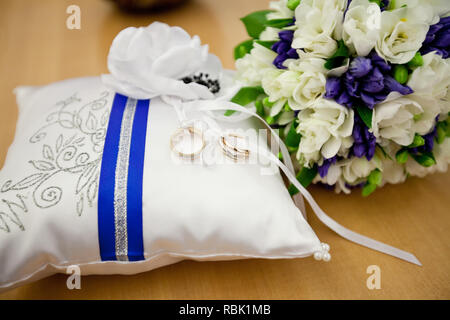 Una coppia di anelli di nozze intrecciate con un nastro bianco su un cuscino bianco. In prossimità di un bouquet di fiori. Foto Stock