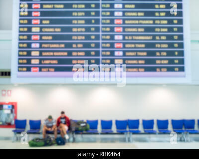 Sfocato l'orario di imbarco degli schermi del monitor e i passeggeri seduti su sedie blu in sala airport terminal area interna per arrivo il passeggero. Foto Stock