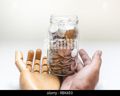 Le monete in un barattolo di vetro a portata di mano in mano in legno isolato su sfondo bianco. Donazione e risparmio di denaro concetto. Foto Stock