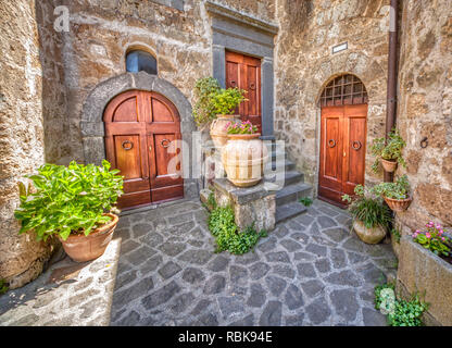 Fotografia di un cortile medievale in Toscana. Tre weathered, usurato in legno marrone di porte con fiori che sbocciano in vasi di argilla posati su un ciottolo pa Foto Stock