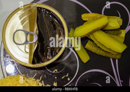 Un piatto lay vista dall'alto di un bianco e nero di taglio di vetro stampato con una lattina aperta di spratti in olio, tritato verdi salati cetrioli e formaggio grattugiato Foto Stock