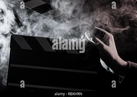 Close-up su un clapperboard aperto a portata di mano prima di iniziare le riprese di un film con il fumo bianco intorno a monocromatico con retroilluminazione su un isolato nero indietro Foto Stock