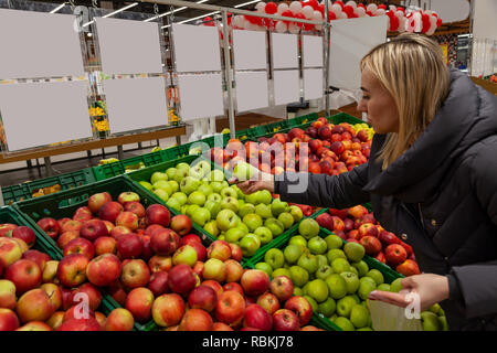 Una donna bionda in un supermercato compra le mele, appoggia attraverso diversi tipi di mele che sono splendidamente esposte sugli scaffali a prendere, arresto Foto Stock