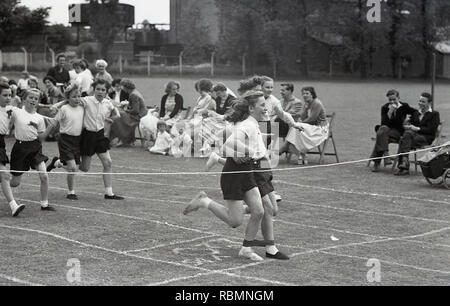anni '1950, storico. Giornata dello sport, bambini delle scuole primarie che partecipano a una gara a tre zampe, Inghilterra, Regno Unito. Due ragazze con un nastro intorno alle gambe che stanno per attraversare la corda finale in una divertente gara sportiva osservata dai genitori dei bambini seduti sul campo d'erba. Foto Stock