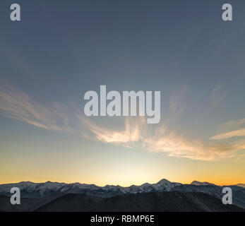 Montagna panorama invernale paesaggio all'alba. Ripide montagne coperte di neve e colline boscose con foreste di abete rosso, sollevamento del sole luminoso su trasparente blu Foto Stock