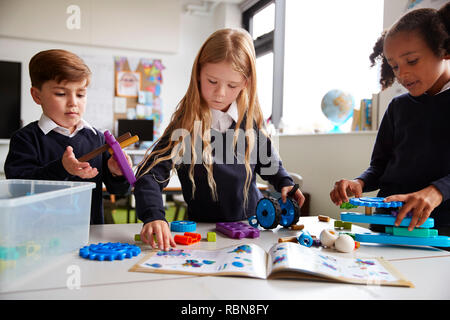 Tre bambini della scuola primaria a lavorare insieme, a seguito di un libretto di istruzioni e utilizzando blocchi di costruzione in una classe, vista frontale, close up Foto Stock