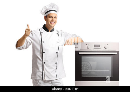 Sorridente maschio giovane chef in piedi accanto ad un forno e dando pollice in alto isolato su sfondo bianco Foto Stock