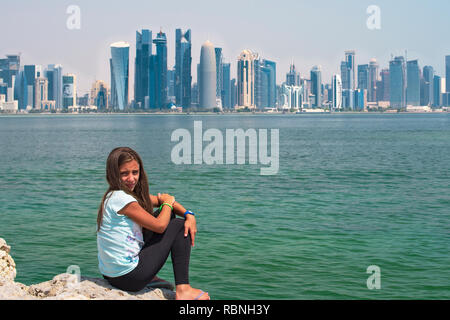 Bambino ragazza seduta sulla Corniche Broadway e guardando a Doha dello Skyline. Il Qatar. Foto Stock