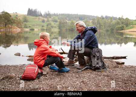 Nonno e nipote seduto sulla riva di un lago insieme sorridente, vicino Lake District, REGNO UNITO Foto Stock