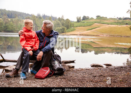 Nipote seduta sul suo nonno del ginocchio sulla riva di un lago sorridente, Lake District, REGNO UNITO Foto Stock