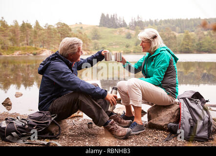 Coppia senior seduta da un lago di bere il caffè durante la vacanza in campeggio fare un brindisi con loro tazze, Lake District, REGNO UNITO Foto Stock